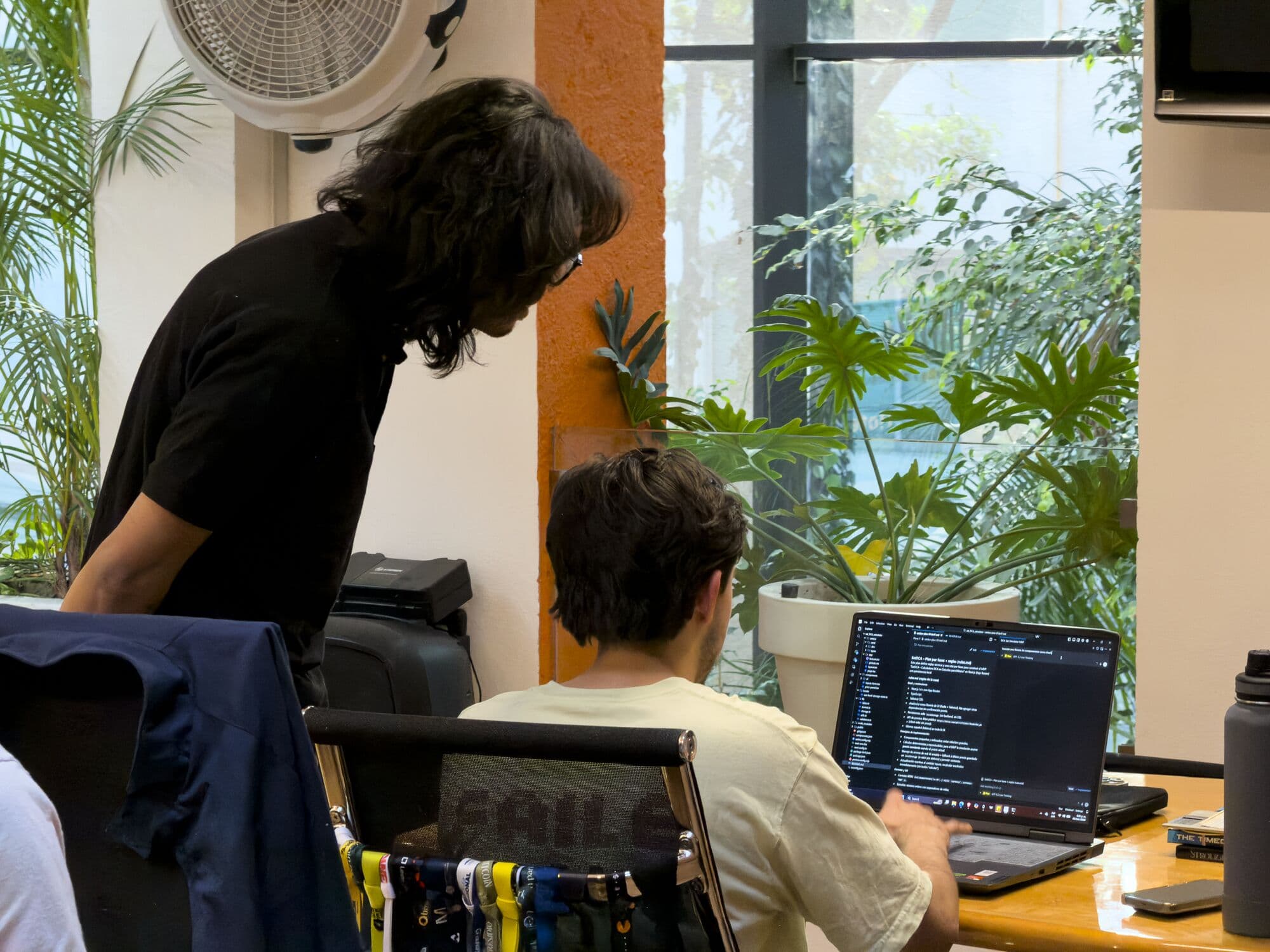 Dos personas revisando código en un escritorio junto a la ventana