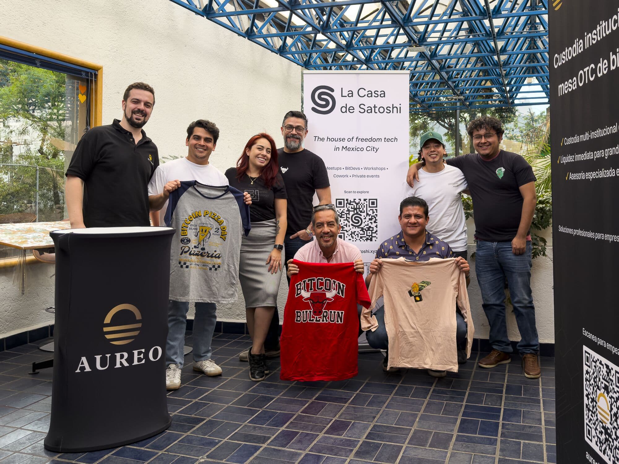 Hackathon participants holding up T-shirts in front of the La Casa de Satoshi banner
